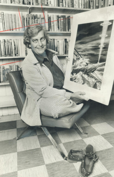 Judith Merril crossed leg on chair holding sci fi art in a small library