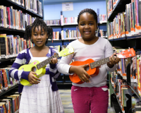 Two children are holding ukuleles in the stacks of a library branch.