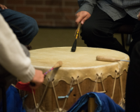 Three drum sticks are hitting a drum. The drum sticks are being held by the hands of different people sitting around the drum.