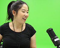 A woman is singing into a microphone that is one or two feet away from her. She is wearing a black shirt and in front of a green screen.