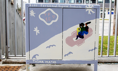 Cabinet outside the branch with painted art of two girls sliding down a snowy hill on a menstrual pad with text that reads: “free personal hygiene products” and “Tasha Matar.”